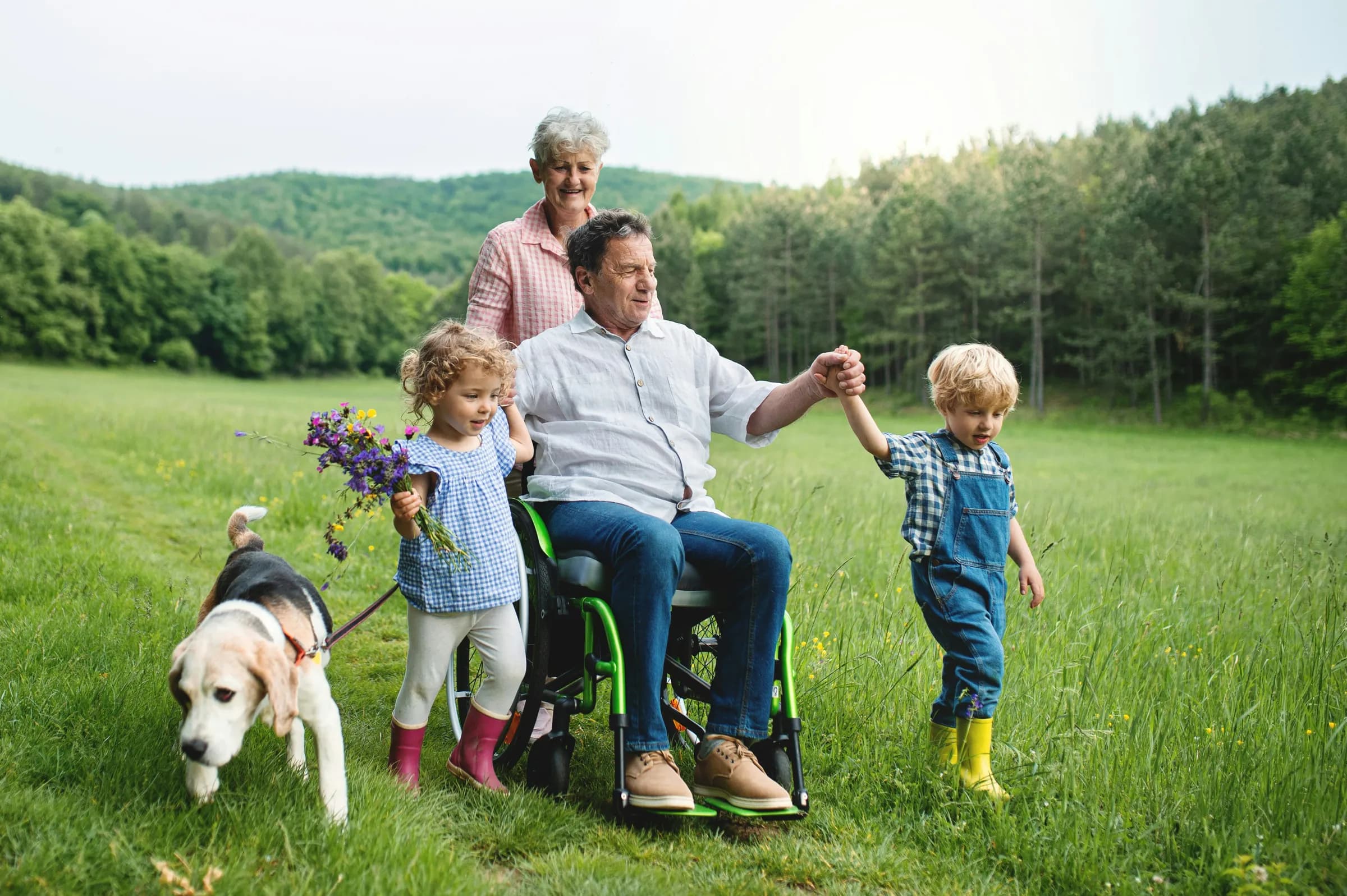 Un grand-père en fauteuil roulant se promène dans un pré avec ses petits-enfants et leur chien