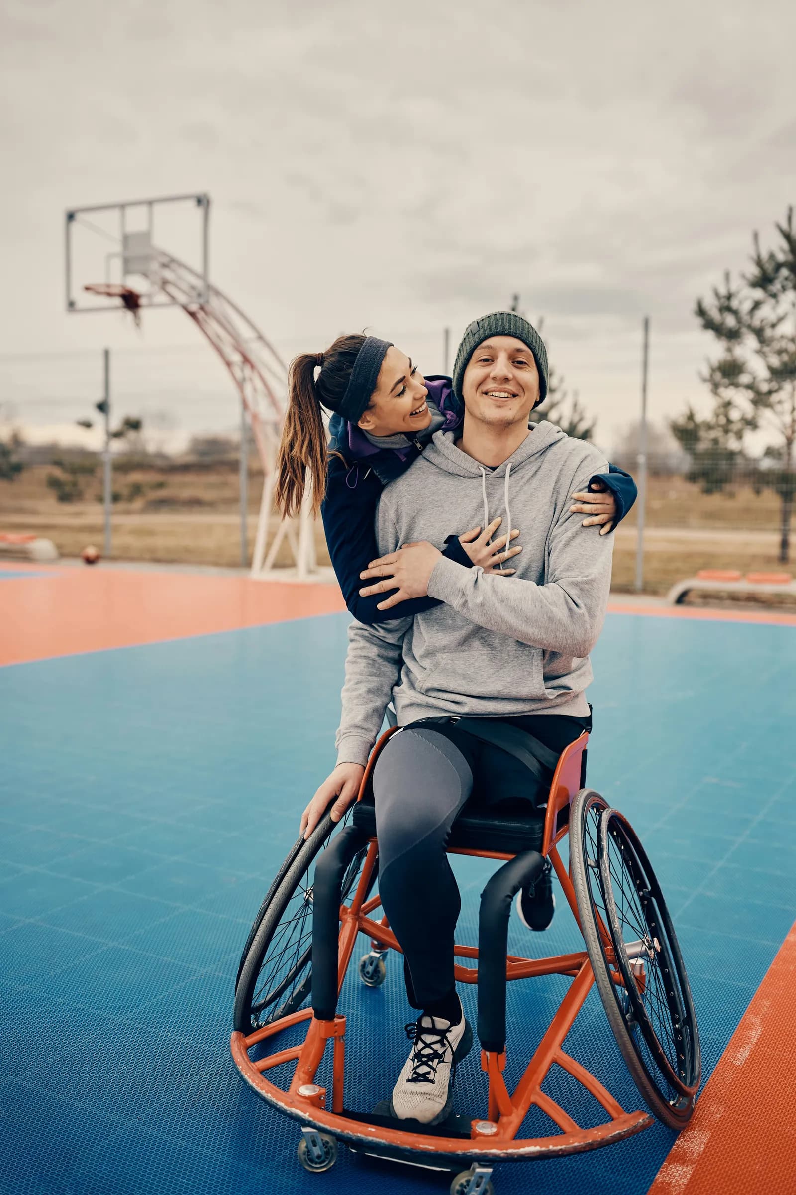 Jeune homme en fauteuil roulant de sport enlacé par sa compagne sur un terrain de basket